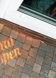 Auf dem gepflasterten Boden vor einem Gebäude ist in oranger Farbe das Wort „Heimatshoppen“ aufgesprüht. Die Aufnahme zeigt einen Ausschnitt des Bodens in der Nähe einer Tür mit Fußmatte.