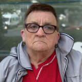 Eine Frau mit Brille und grauer Jacke blickt in die Kamera. Im Hintergrund ist ein Fenster mit Reflektionen zu sehen.