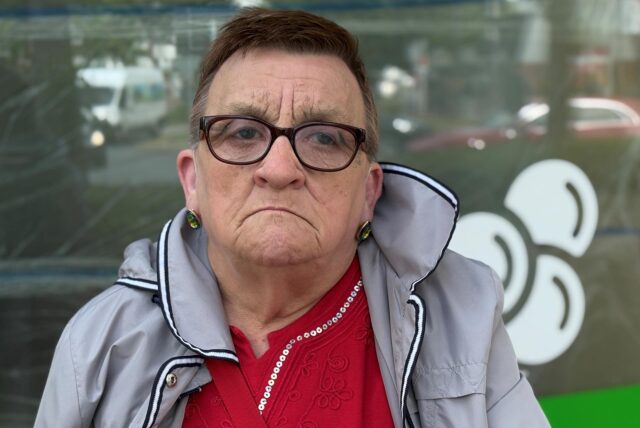 Eine Frau mit Brille und grauer Jacke blickt in die Kamera. Im Hintergrund ist ein Fenster mit Reflektionen zu sehen.