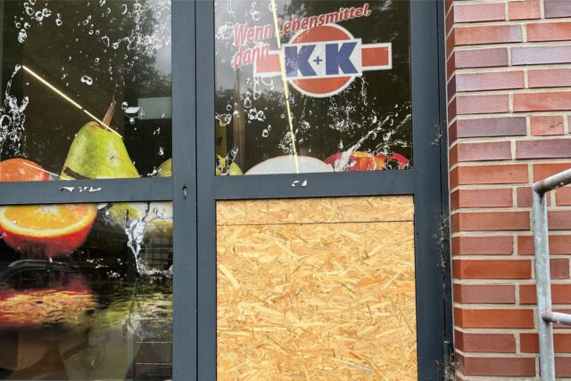 Die Aufnahme zeigt die Eingangstür eines Ladens, die teilweise mit einer Holzplatte verbarrikadiert ist.