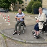 Zwei Senioren sitzen auf Rollatoren vor einem Parkplatz in Marl.