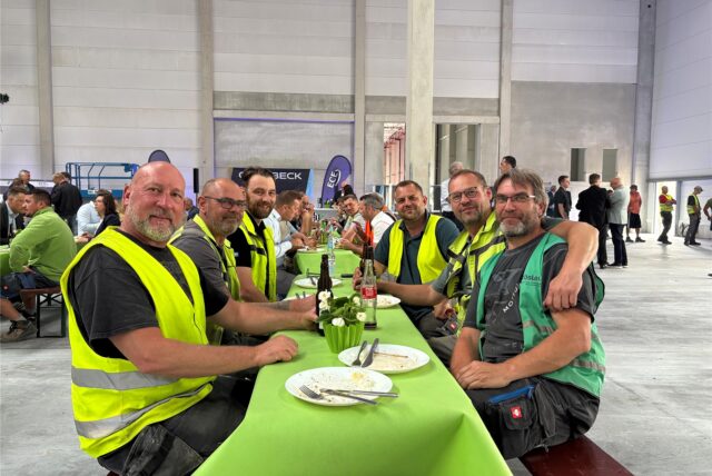 Eine Gruppe von Bauarbeitern posiert für ein Foto an einem mit einer grünen Tischdecke bedeckten Tisch. Sie sitzen auf einer Bank in der neuen Thalia-Halle.