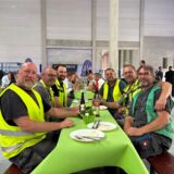 Eine Gruppe von Bauarbeitern posiert für ein Foto an einem mit einer grünen Tischdecke bedeckten Tisch. Sie sitzen auf einer Bank in der neuen Thalia-Halle.