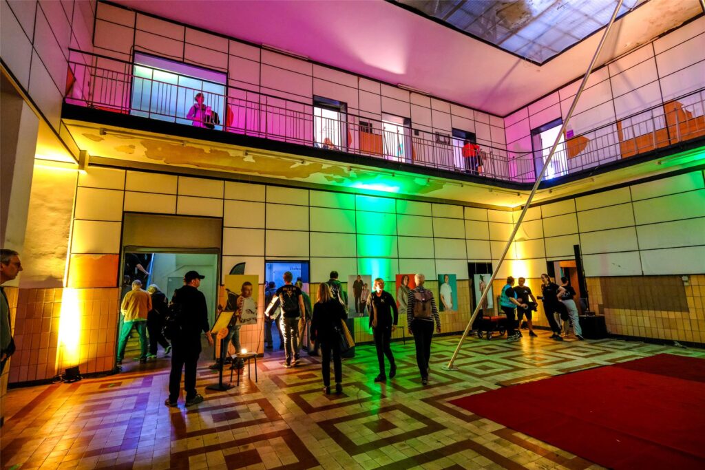 Blick in die bunt beleuchtete Lohn- und Lichthalle der Neuen Zeche Westerholt während der ExtraSchicht 2024, in der einige Besucher Kunstwerke anschauen.