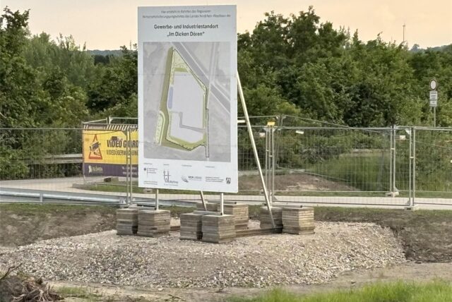 Ein Schild zeigt die geplante Entwicklung eines Gewerbegebiets. Dahinter sind Zäune und einige Sträucher zu sehen.