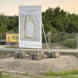 Ein Schild zeigt die geplante Entwicklung eines Gewerbegebiets. Dahinter sind Zäune und einige Sträucher zu sehen.