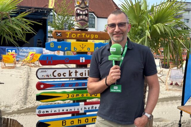 Reporter Jürgen Bröker steht vor einer mit Sand aufgeschütteten Fläche und einem bunten Schild.