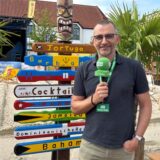 Reporter Jürgen Bröker steht vor einer mit Sand aufgeschütteten Fläche und einem bunten Schild.