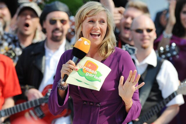 Andrea Kiewel mit ZDF-Mikrofon und Moderationskarten, während sie den ZDF-Fernsehgarten moderiert. Im Hintergrund ist eine Musikgruppe zu sehen.