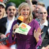 Andrea Kiewel mit ZDF-Mikrofon und Moderationskarten, während sie den ZDF-Fernsehgarten moderiert. Im Hintergrund ist eine Musikgruppe zu sehen.