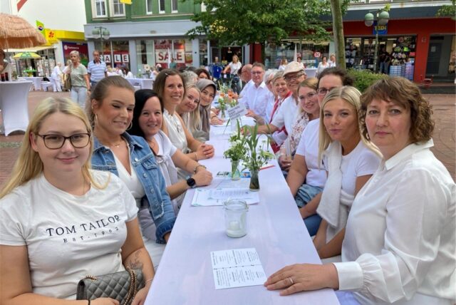 Eine Gruppe von Personen sitzt an einem langen Tisch im Freien, der mit einer weißen Tischdecke bedeckt ist. Die meisten Anwesenden tragen weiße Kleidung.