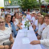 Eine Gruppe von Personen sitzt an einem langen Tisch im Freien, der mit einer weißen Tischdecke bedeckt ist. Die meisten Anwesenden tragen weiße Kleidung.