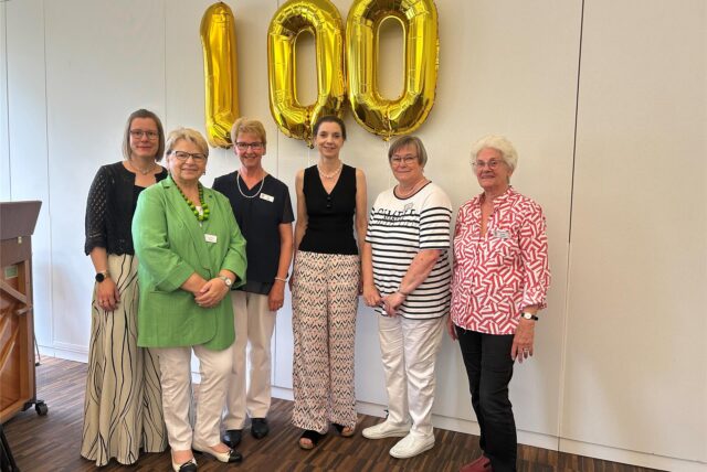 Sechs Frauen stehen vor goldenen Ballons, die eine 100 zeigen