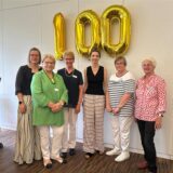 Sechs Frauen stehen vor goldenen Ballons, die eine 100 zeigen