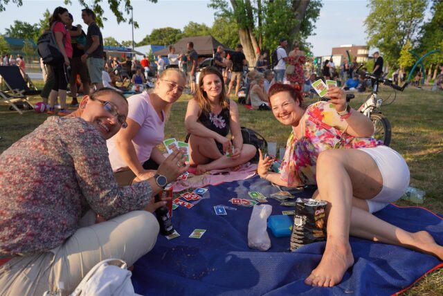 Eine Gruppe von vier Frauen sitzt auf einer blauen Decke im Freien und spielt Skip-Bo. Im Hintergrund sind weitere Personen und ein Fahrrad zu sehen.