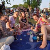 Eine Gruppe von vier Frauen sitzt auf einer blauen Decke im Freien und spielt Skip-Bo. Im Hintergrund sind weitere Personen und ein Fahrrad zu sehen.