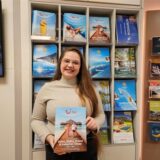 Michelle Doctor zeigt einen Reisekatalog in die Kamera und steht vor einem Regal, in dem sich noch weitere Kataloge befinden.
