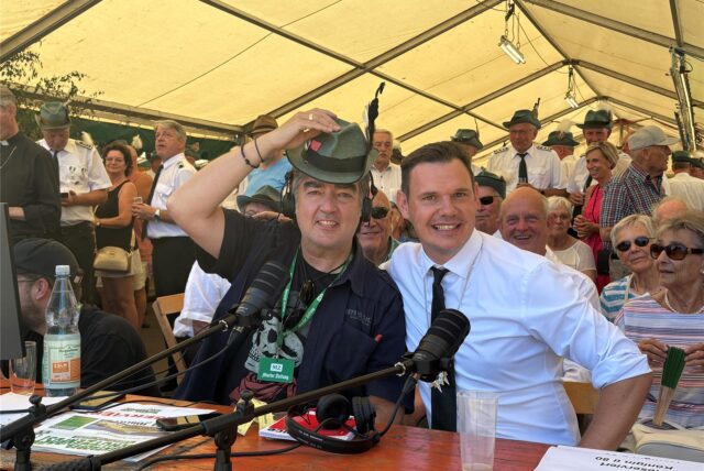 Live vom Königsschießen in Marl-Frentrop: MZ-Redakteur Patrick Köllner (li.) und Christoph Winkel von den Frentroper Schützen sitzen am Kommentatoren-Tisch und lächeln in die Kamera.