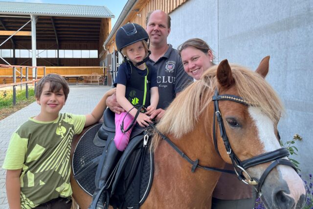 Auf dem Bild ist eine Familie mit zwei Kindern und einem Pferd zu sehen. Das jüngere Kind sitzt mit Reitausrüstung auf dem Pferd, während die Eltern und das ältere Kind daneben stehen.