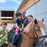Auf dem Bild ist eine Familie mit zwei Kindern und einem Pferd zu sehen. Das jüngere Kind sitzt mit Reitausrüstung auf dem Pferd, während die Eltern und das ältere Kind daneben stehen.