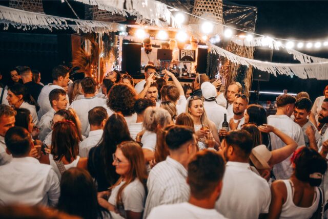 Party-Gäste in weißer Kleidung tanzen, im Hintergrund legt ein DJ auf.