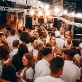 Party-Gäste in weißer Kleidung tanzen, im Hintergrund legt ein DJ auf.
