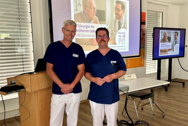 Dr. Christian Kühne, Chefarzt für Chirurgie am EvK Castrop-Rauxel, und Olaf Trenke, Oberarzt der Klinik für Anästhesie, stehen vor einer Leinwand, auf der eine Präsentation zu sehen ist.
