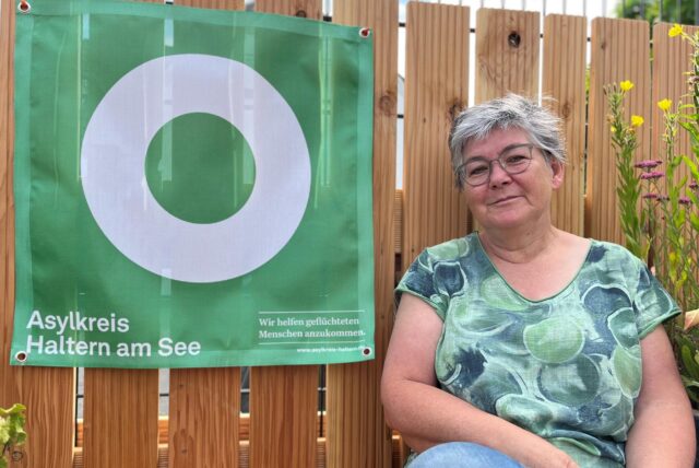 Ute Erler vom Asylkreis Haltern sitzt im Garten der Geschäftsstelle auf einer Bank. Neben ihr hängt das Logo des Asylkreises.