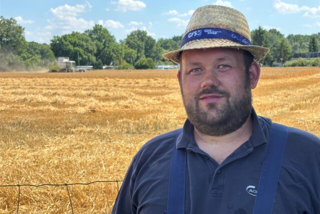 Ein Mann mit Bart und Strohhut steht vor einem Feld. Darauf fährt ein Mähdrescher. Im Hintergrund sind Bäume und blauer Himmel zu sehen.