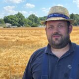 Ein Mann mit Bart und Strohhut steht vor einem Feld. Darauf fährt ein Mähdrescher. Im Hintergrund sind Bäume und blauer Himmel zu sehen.