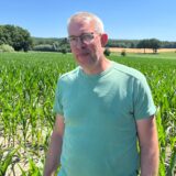 Ein Mann mit Brille steht vor einem Feld junger Maispflanzen unter blauem Himmel. Im Hintergrund sind Bäume und weitere Felder zu sehen.