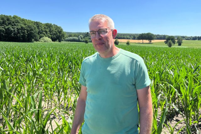Ein Mann mit Brille steht vor einem Feld junger Maispflanzen unter blauem Himmel. Im Hintergrund sind Bäume und weitere Felder zu sehen.