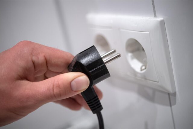 Eine Hand hält einen schwarzen Stecker in Richtung einer weißen Doppelsteckdose an einer weißen Wand. Der Stecker ist noch nicht eingesteckt.