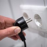 Eine Hand hält einen schwarzen Stecker in Richtung einer weißen Doppelsteckdose an einer weißen Wand. Der Stecker ist noch nicht eingesteckt.