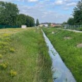Eine malerische Landschaft zeigt einen kleinen Flusslauf, der von üppigem Grün und Bäumen gesäumt ist. Im Hintergrund sind Wohnhäuser und ein bewölkter Himmel sichtbar. Daneben ist das Foto eines Mannes zu sehen, der ein weißes Hemd und eine graue Anzugsjacke trägt.