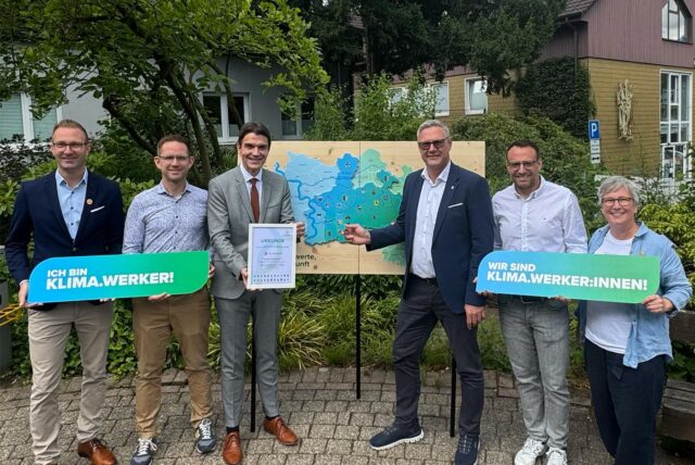 ine Gruppe von sechs Personen steht vor einer Tafel mit der Karte des Kreises Recklinghausen. Die Personen halten Schilder mit der Aufschrift „Ich bin Klima.Werker!“ und „Wir sind Klima.Werker:innen!“.
