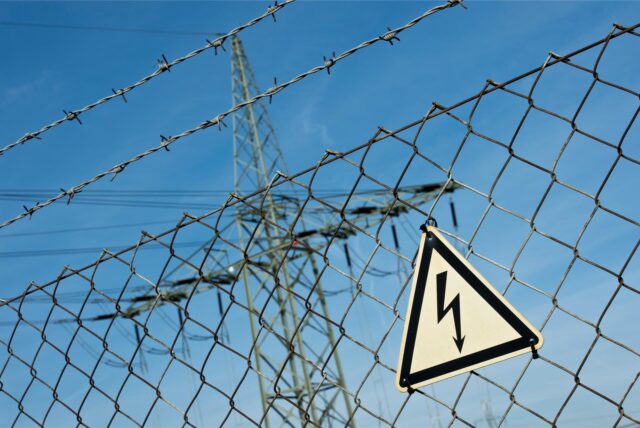 Ein Gefahrenschild für Elektrizität ist vor einer Stromtrasse zu sehen.