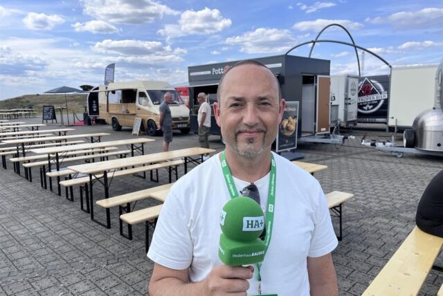 HA-Redakteur André Przybyl steht mit einem grünen Mikrofon in der Hand vor den Foodtrucks und Tischen des Street-Food-Festivals auf der Halde Hoheward in Herten.