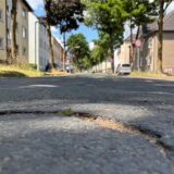 Eine Nahaufnahme zeigt einen Riss im Asphalt einer Straße, aus dem ein kleiner grüner Trieb sprießt. Im Hintergrund sind Wohnhäuser, geparkte Autos und ein Geschwindigkeitsbegrenzungsschild zu erkennen.