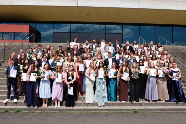 Die Abiturienten 2025 am Herwig-Blankertz-Berufskolleg stehen mit ihren Zeugnissen auf der Treppe am Campus Vest in Recklinghausen.