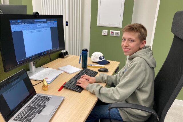 Ein junger Mann sitzt an einem Schreibtisch und tippt auf einer Computertastatur. Auf dem Tisch befinden sich außerdem ein Laptop, ein Monitor, eine Wasserflasche und eine Kappe mit einer Aufschrift.