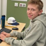 Ein junger Mann sitzt an einem Schreibtisch und tippt auf einer Computertastatur. Auf dem Tisch befinden sich außerdem ein Laptop, ein Monitor, eine Wasserflasche und eine Kappe mit einer Aufschrift.