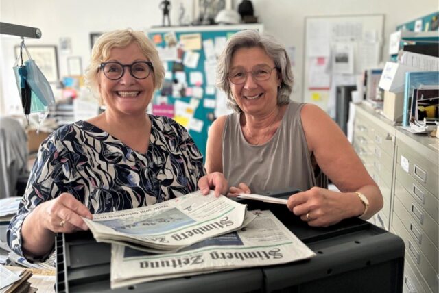 Zwei Frauen stehen in einem Büro. Vor sich haben sie Zeitungen. Beide tragen eine Brille.