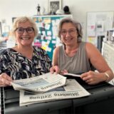 Zwei Frauen stehen in einem Büro. Vor sich haben sie Zeitungen. Beide tragen eine Brille.