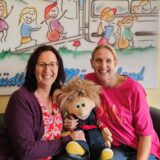 Die Leiterinnen des Kinder- und Jugendhospizes in Haltern Katja Büscher-Mönig und Yvonne Steinbuße halten zusammen eine Puppe fest und lachen in die Kamera.