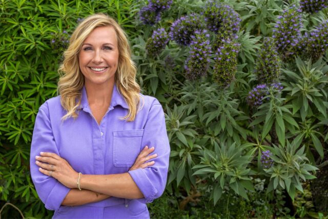 Andrea Kiewel, Moderatorin des ZDF-Fernsehgarten steht in einem lila Hosenanzug vor einer Hecke.