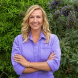 Andrea Kiewel, Moderatorin des ZDF-Fernsehgarten steht in einem lila Hosenanzug vor einer Hecke.