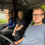 Drei Personen sitzen in der Fahrerkabine eines LKW der Marke DAF. Ein Junge mit Brille sitzt am Steuer, während zwei Mädchen auf den Passagiersitzen Platz genommen haben.