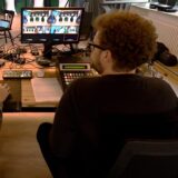 Zwei Männer sitzen mit dem Rücken zur Kamera vor Technikpulten, im Hintergrund stehen Menschen an Stehtischen.