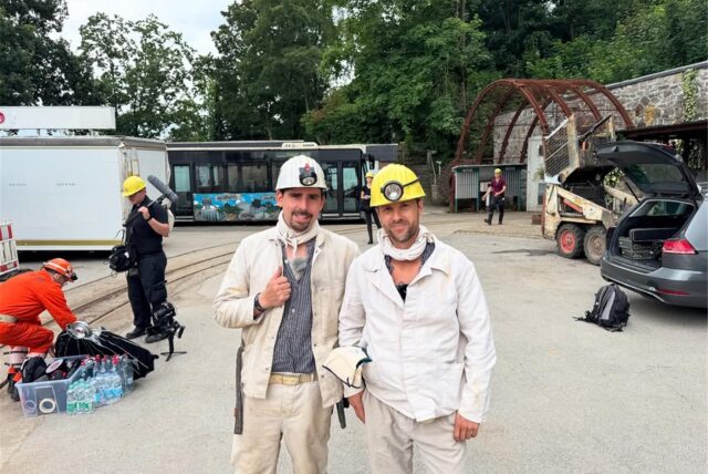 Marcel Grigull und Mirko Drotschmann posieren für ein Bild in einem Bergbauoutfit. Im Hintergrund ist ein Kamerateam zu erkennen und das Gelände des Trainingbergwerks Recklinghausen.